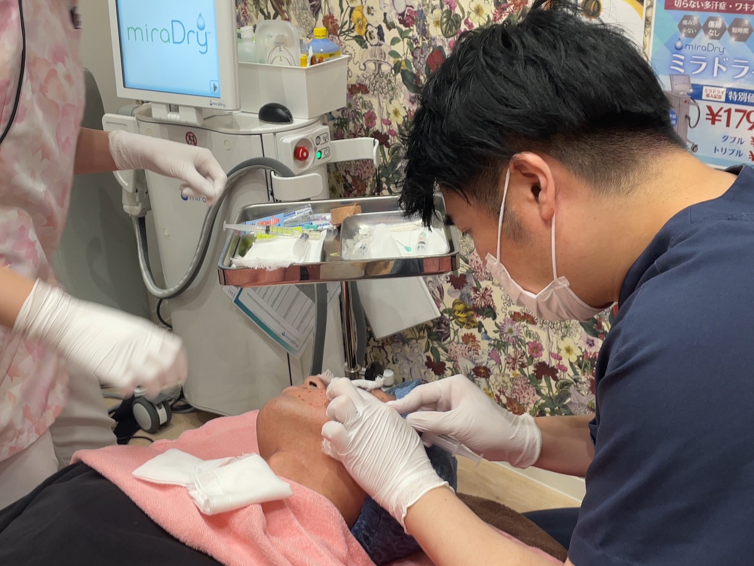 横たわる人の頬に器具を当てる施術の様子 マスクと手袋を着けた人物が、ベッドに横たわる人の頬に器具を当てている。そばには手袋を着けた別の人物がおり、器具を並べたトレーと画面付きの機器が置かれている。